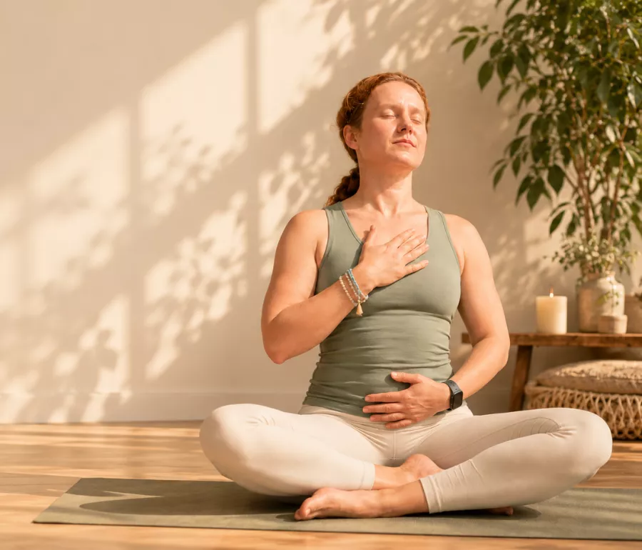 Vrouw in kleermakerszit op een yogamat, ogen dicht, met een hand op haar borst en een hand op haar buik